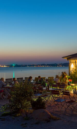 Menschen sitzen abends im Außenbereich eines Cafés am Wasser mit Blick auf die beleuchtete Stadt Kiel
